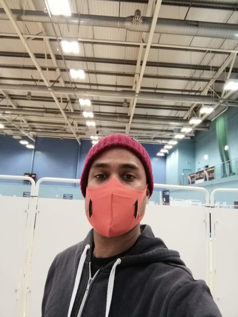 Photo of me standing at the entrance to the vaccination centre to get my first dose of Pfizer covid-19 vaccination. I'm wearing a beige beanie hat, navy hoodie and orange U-mask. 